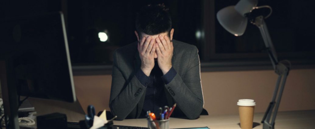 Tired young man in suit and tie is working on computer in office at night sitting at desk in dark room, looking at screen and typing.