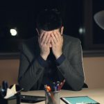 Tired young man in suit and tie is working on computer in office at night sitting at desk in dark room, looking at screen and typing.